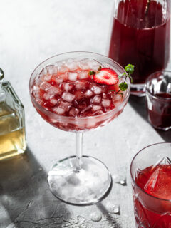 A stemmed coup glass filled with a red drink and ice, in front of a glass carafe filled with strawberry shrub.
