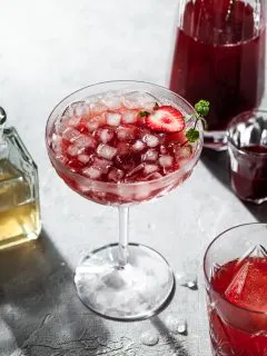 A stemmed coup glass filled with a red drink and ice, in front of a glass carafe filled with strawberry shrub.