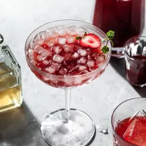 A stemmed coup glass filled with a red drink and ice, in front of a glass carafe filled with strawberry shrub.