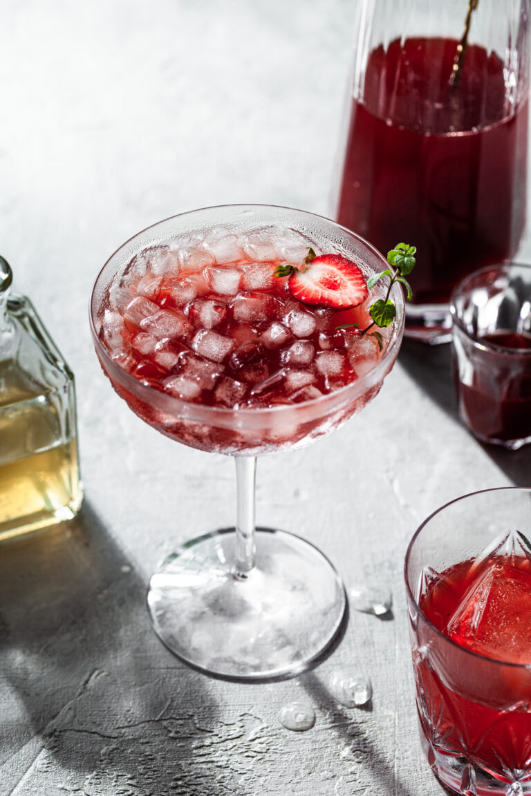 A stemmed coup glass filled with a red drink and ice, in front of a glass carafe filled with strawberry shrub.