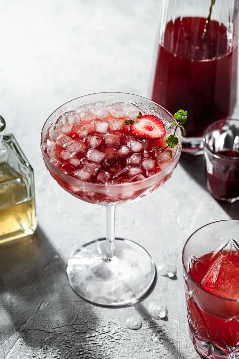 A stemmed coup glass filled with a red drink and ice, in front of a glass carafe filled with strawberry shrub.