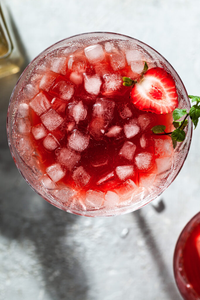 View looking down into a glass filled with crushed ice in a red drink (strawberry shrub) garnished with a strawberry and a sprig of mint. 