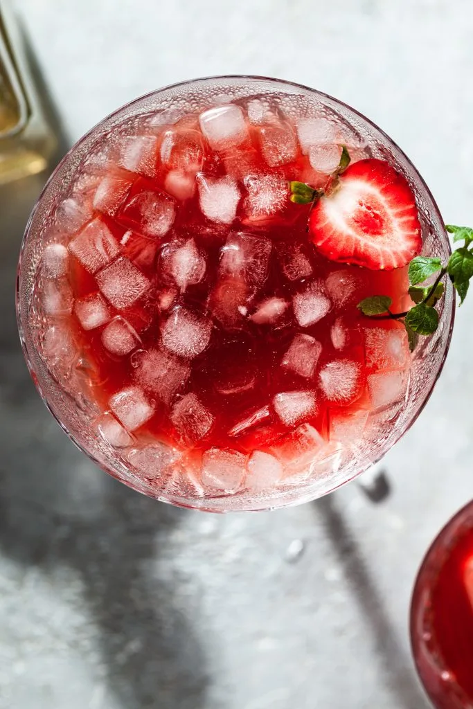 View looking down into a glass filled with crushed ice in a red drink (strawberry shrub) garnished with a strawberry and a sprig of mint.