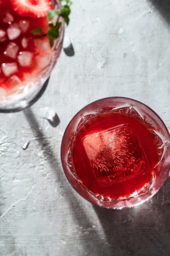 View looking down into a crystal glass filled with a red beverage and a square ice cube, on a grey counter in bright sunlight.