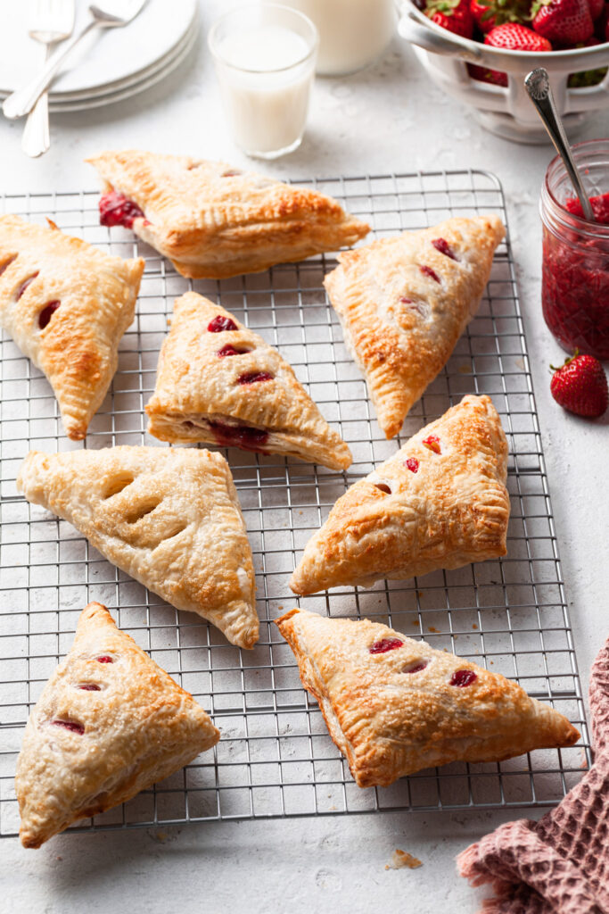 8 turnovers on a wire cooling rack, with a basket of strawberries in the background with a jar of red berry preserves.