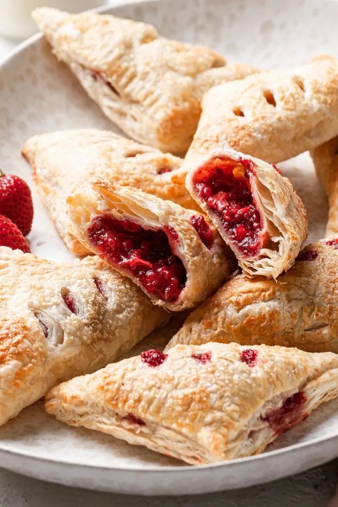 Close up of a hand pie sliced open, displaying the red filling. The sliced hand pie sits on top of other pastries in a curved cream platter. 