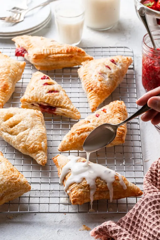 White glaze being drizzled from a silver spoon onto a turnover on a wire rack. On the same wire rack are 7 other pastries.