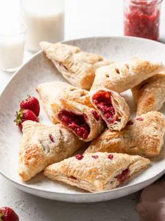 Strawberry turnovers in a curved cream colored platter. One turnover has been cut in half to reveal the red fruit center.