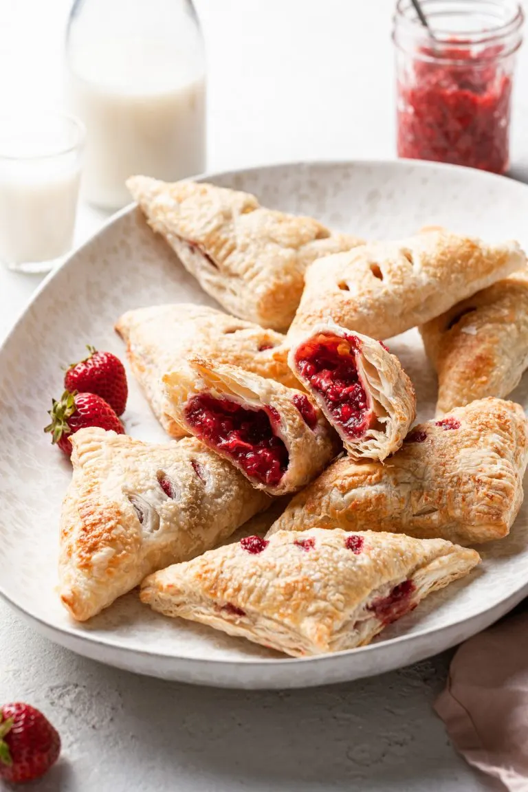 Strawberry turnovers in a curved cream colored platter. One turnover has been cut in half to reveal the red fruit center.