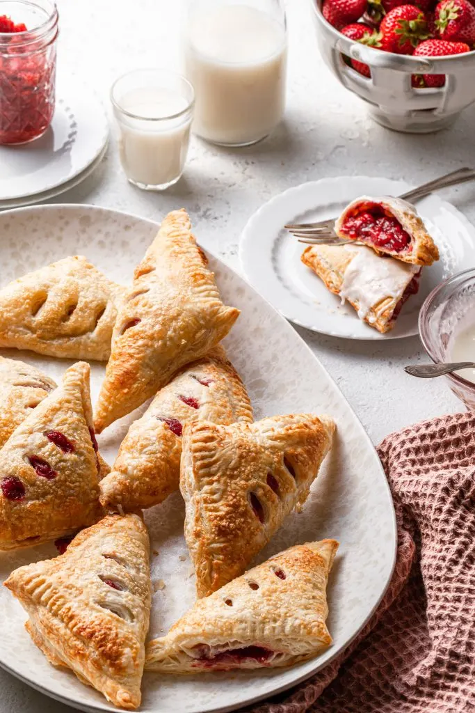 A tray filled with pasties, in the background is a white plate with a sliced open pasty covered in white glaze, and a couple of glasses of milk. 