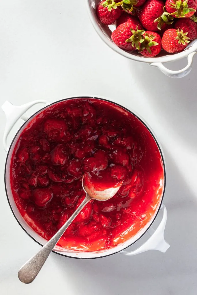 A white enameled pot filled with strawberry jam, beside a white porcelain berry basket filled with fresh berries.  