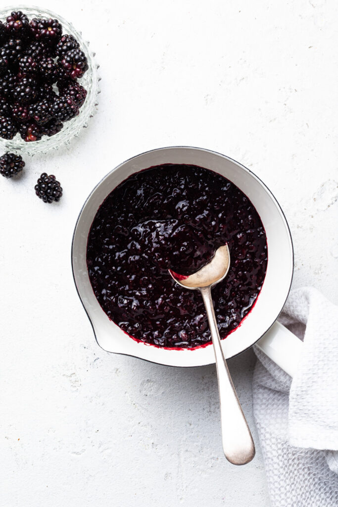 A white enamel pot filled with cooked purple berries, and a silver spoon. 