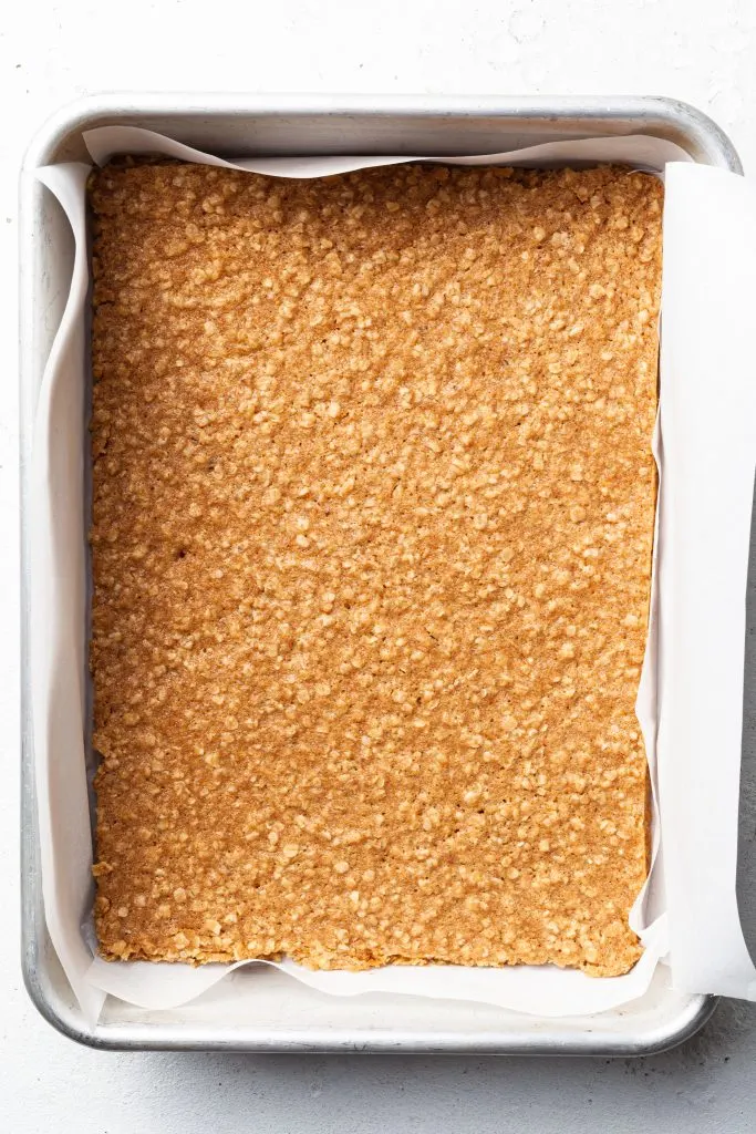 A rectangular baking tin lined with white parchment paper and filled with a baked oatmeal cookie bar.  