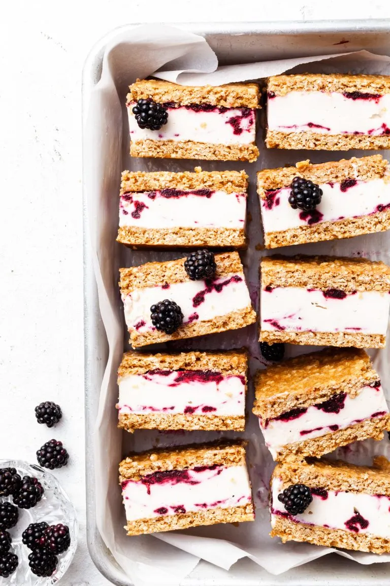 Baking tin lined with white parchment, filled with ice cream sandwiches that contain and purple swirl through the white ice cream layer.