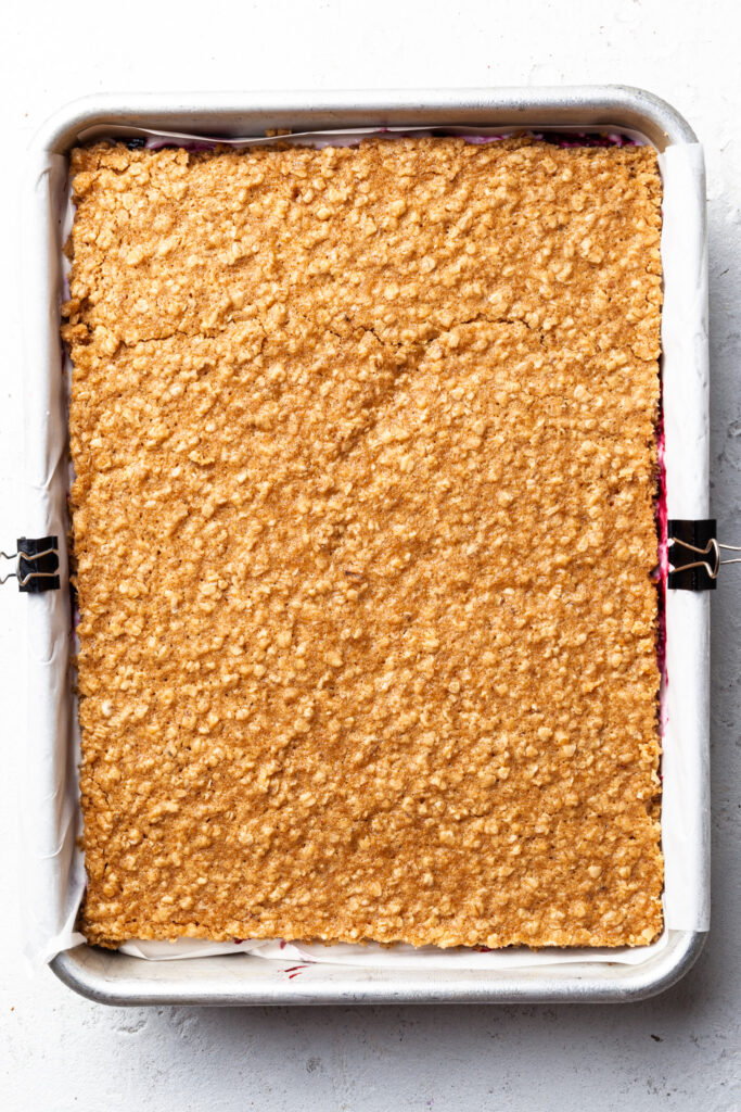 A rectangular baking tin lined with parchment and filled with an assembled dessert. Oatmeal cookie slab on top, with berry jam peeking out the sides. 