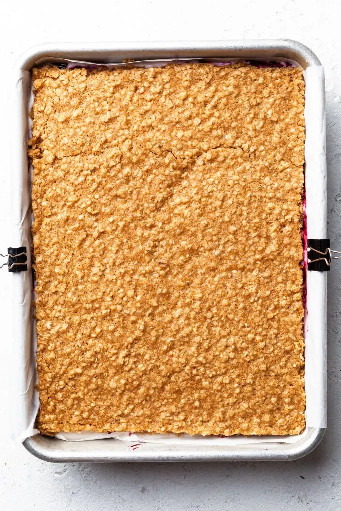 A rectangular baking tin lined with parchment and filled with an assembled dessert. Oatmeal cookie slab on top, with berry jam peeking out the sides. 