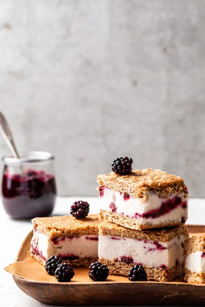 4 ice cream sandwiches on a wooden plate, 2 are stacked. Fresh blackberries are scattered about, and a jar of jam sits in the background. 