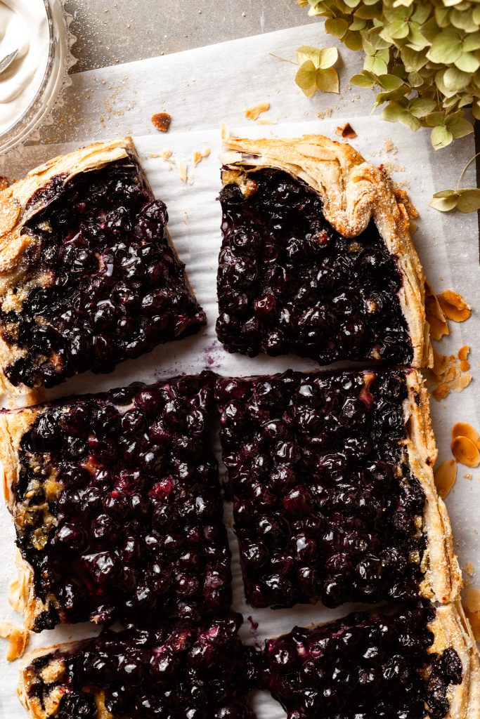 A berry galette sliced on a piece of parchment paper beside a bowl of whipped cream. 