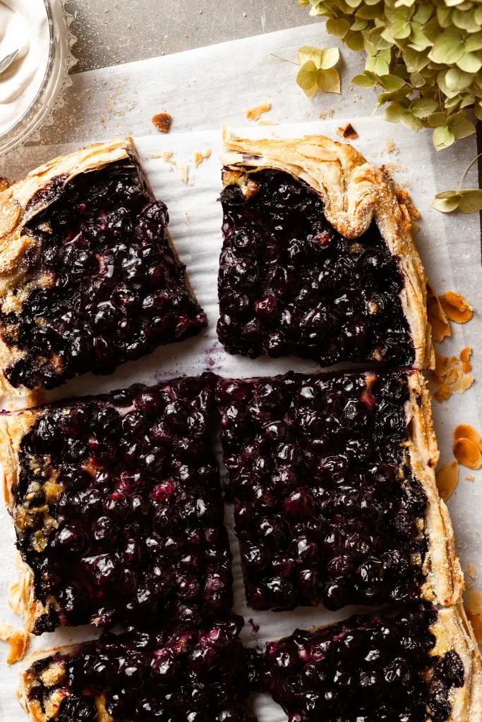A berry galette sliced on a piece of parchment paper beside a bowl of whipped cream. 