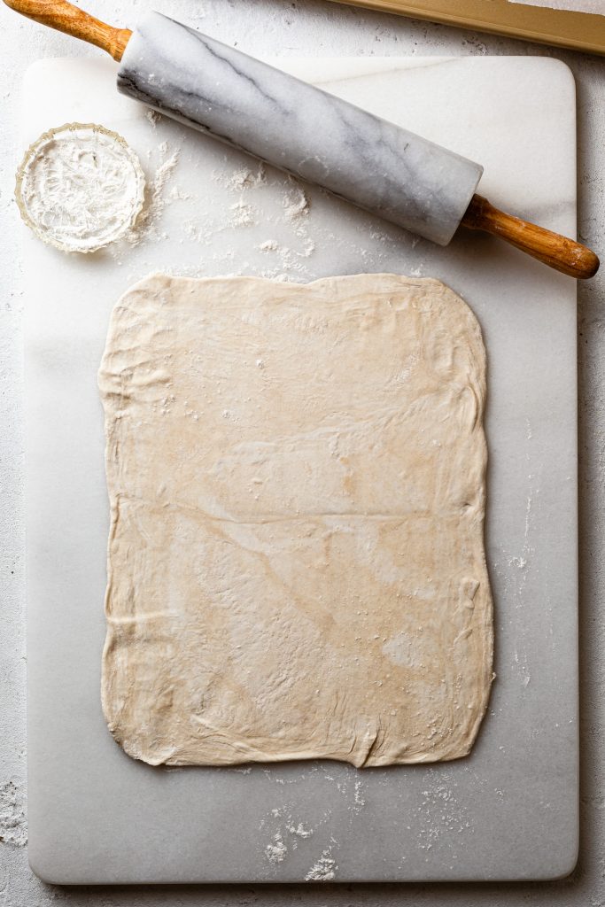 Pastry rolled out into the shape of a rectangle, on a white marble board next to a marble rolling pin.