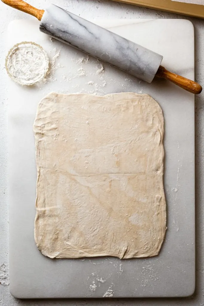 Pastry rolled out into the shape of a rectangle, on a white marble board next to a marble rolling pin.