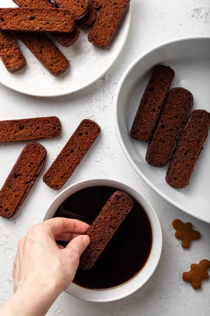A hand dipping a cookie into a bowl of coffee, beside a baking tray partly lined with already dipped cookies.