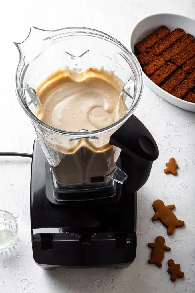 A blender filled with a creamy mixture, and a dish in the background partly lined with gingerbread cookies. 