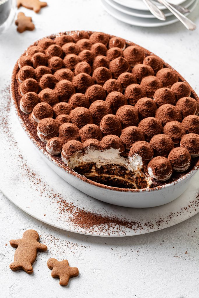 An oval dish filled with tiramisu with a serving missing, and gingerbread cookies laying on the same white counter. 