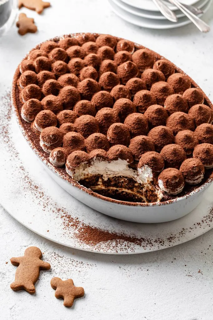 An oval dish filled with tiramisu with a serving missing, and gingerbread cookies laying on the same white counter. 