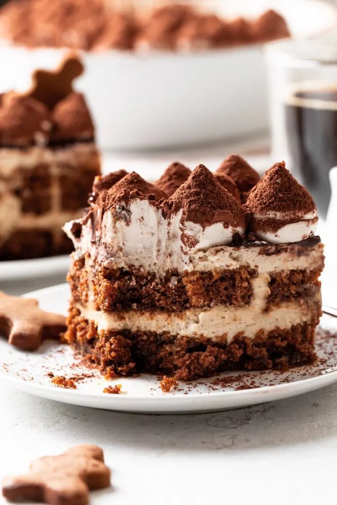 Close up shot of a piece of tiramisu served on a white plate, with a piece missing. Gingerbread cookies are on the same white counter, and the serving plate. 
