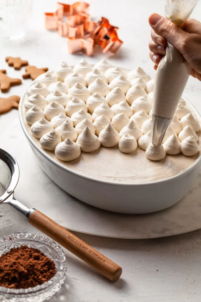 A hand squeezing a pastry bag filling with whipped cream, creating dollops on top of a dish of tiramisu. 