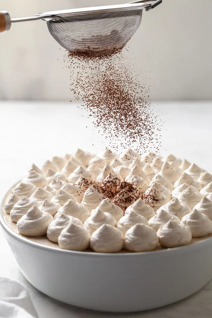 A sieve of cocoa powder being shaken over a white dish of tiramisu. 