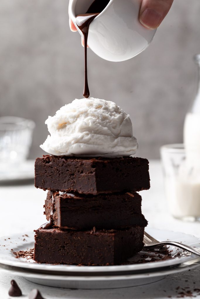 A stack of three brownies topped with a scoop of vanilla ice cream and a drop of chocolate sauce pouring from a small white jug above.