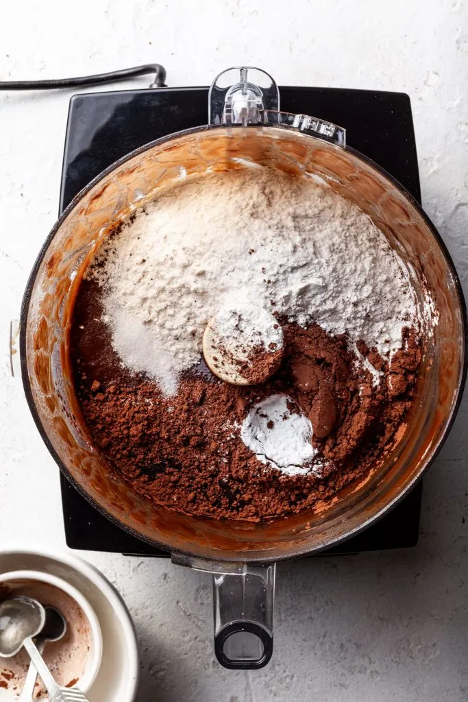 A food processor filled with a liquid chocolate mixture, and freshly added not yet blended dry ingredients. 
